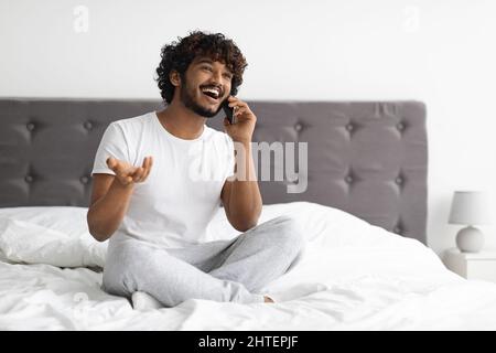 Un homme hindou positif assis au lit, parlant au téléphone Banque D'Images