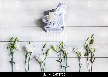 Boîte cadeau tendance enveloppée dans un tissu de soie de Furoshiki technique, les fleurs blanches Eustoma ou Lisianthus se trouvent dans une rangée au fond sur le dos en bois clair Banque D'Images