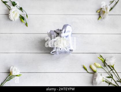 Boîte cadeau tendance enveloppée dans un tissu de soie de Furoshiki technique, fleurs blanches Eustoma ou Lisianthu dans les coins sur fond de bois clair vue du dessus f Banque D'Images