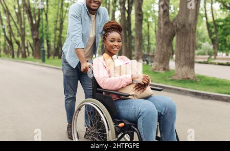 Belle jeune femme noire en fauteuil roulant tenant un sac de papier avec de la nourriture après avoir fait du shopping avec son petit ami à l'extérieur Banque D'Images