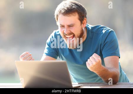 Un homme enthousiaste qui vérifie le contenu d'un ordinateur portable et célèbre les bonnes nouvelles en plein air Banque D'Images