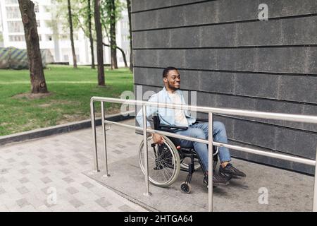 Concept d'environnement accessible pour les personnes ayant des besoins spéciaux. Jeune homme noir positif en fauteuil roulant utilisant la rampe à l'extérieur Banque D'Images