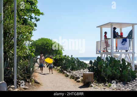 El Tunco, El Salvador - 29 janvier 2022: Surfeurs marchant le long de la plage rocheuse pour trouver des vagues parfaites pour le surf. Sauveteurs debout sur la chaise Banque D'Images