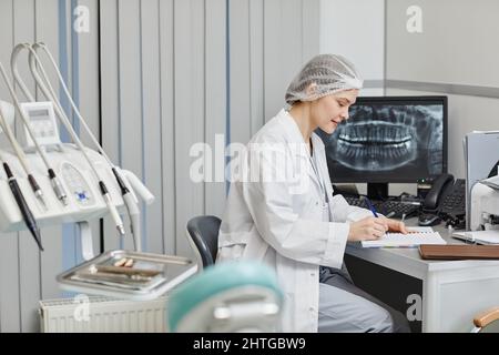 Portrait de jeune femme dentiste en vue latérale écrivant sur une planchette à pince tout en travaillant au bureau de la clinique dentaire, espace de copie Banque D'Images