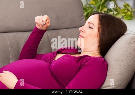 Photo d'une femme enceinte malade avec robe lilas vérifiant la température du thermomètre assis sur un canapé dans le salon dans une maison intérieure Banque D'Images
