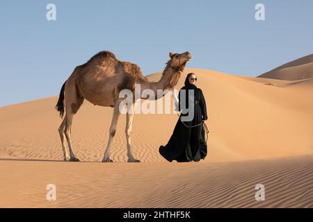 Jeune femme en robe nationale Emirati (abaya) avec un chameau dans les dunes désertiques des quartiers vides. Abu Dhabi, Émirats arabes Unis. Banque D'Images