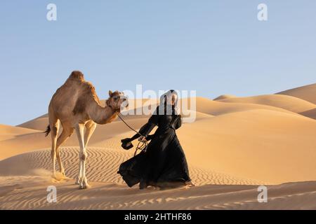 Jeune femme en robe nationale Emirati (abaya) avec un chameau dans les dunes désertiques des quartiers vides. Abu Dhabi, Émirats arabes Unis. Banque D'Images