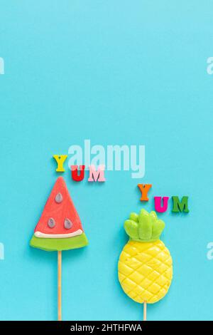 Yum Yum texte coloré, sucettes ananas et pastèque sur bâton sur fond bleu jaune. Modèle Creative Top View pour les vacances ou les fêtes de fin d'année Banque D'Images