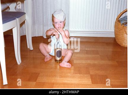 Photographie des années 60 de petit enfant jouant avec une bouteille de verre assise sur un plancher en bois, Suède. Banque D'Images