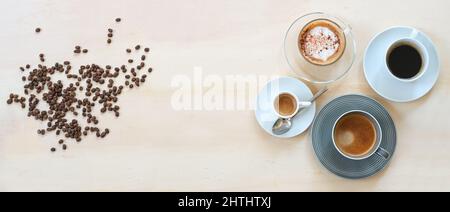 Grains de café rôtis et quatre tasses avec différentes boissons comme, espresso, cappuccino, lungo noir et avec crème sur une table en bois clair, panoramique forma Banque D'Images