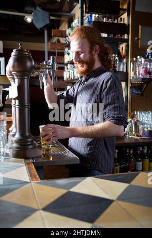 Vue latérale d'un barkeeper mâle optimiste avec des cheveux rouges versant de la bière sur le robinet dans le verre tout en se tenant au comptoir pendant le travail au bar Banque D'Images