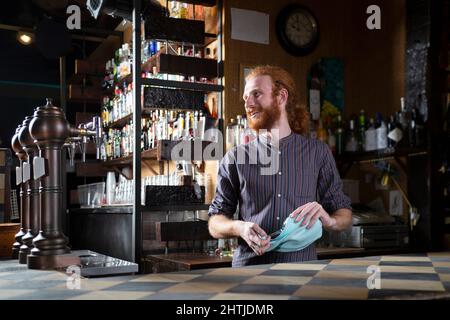 Barman optimiste avec des cheveux rouges essuyant des verres de vin avec une serviette de table tout en se tenant au comptoir dans le bar avec diverses bouteilles d'alcool Banque D'Images