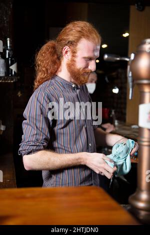 Barman optimiste avec des cheveux rouges essuyant des verres de vin avec une serviette de table tout en se tenant au comptoir dans le bar avec diverses bouteilles d'alcool Banque D'Images