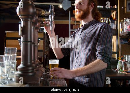 Vue latérale d'un barkeeper court avec cheveux rouges qui verse de la bière sur le robinet dans le verre tout en se tenant au comptoir pendant le travail au bar Banque D'Images