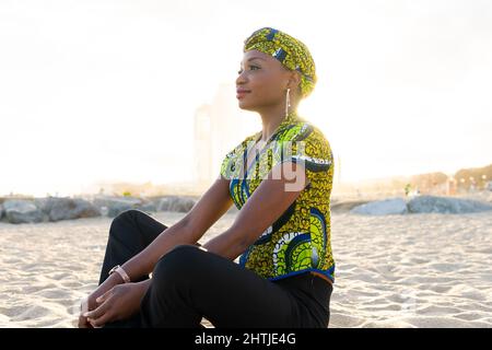 Femme africaine détendue dans un foulard traditionnel souriant et regardant loin tout en étant assise en croix sur la plage de sable et méditant au coucher du soleil Banque D'Images