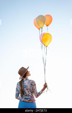 Vue arrière d'une femme inreconnaissable dans un chapeau tenant un tas de ballons colorés tout en se tenant contre le ciel clair Banque D'Images