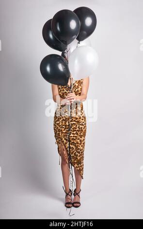 Corps complet de femme méconnue dans une robe élégante couvrant le visage avec un bouquet de ballons debout sur fond blanc tout en célébrant l'anniversaire de St Banque D'Images