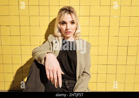 Jeune femme avec une tenue élégante regardant l'appareil photo tout en étant assise près d'un mur carrelé jaune Banque D'Images