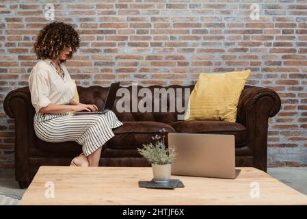 Vue latérale d'une travailleuse concentrée qui navigue sur un netbook tout en étant assise sur un canapé confortable dans un bureau léger et élégant avec un mur de briques Banque D'Images