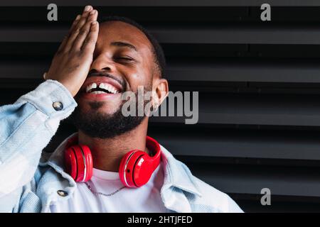 Homme afro-américain à barbe avec casque sans fil rouge moderne couvrant un œil fermé tout en restant debout sur fond noir dans la rue Banque D'Images