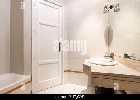 Salle de bains avec meuble-lavabo en marbre crème et lavabo en porcelaine blanche avec miroir en verre sans cadre carré et porte en bois blanc Banque D'Images