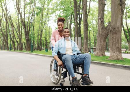 Joyeux jeune couple noir avec une femme charmante et un millennium en fauteuil roulant allant pour marcher au parc Banque D'Images
