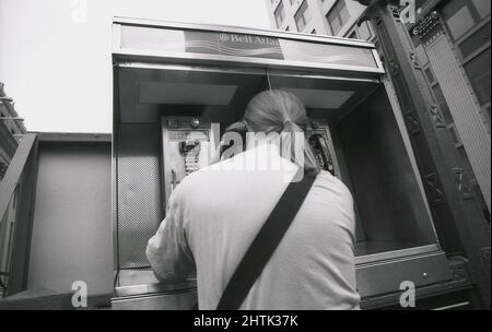 1980s, historique, un homme avec une queue de poney, à une cabine téléphonique ouverte ou non fermée, à l'aide d'un bouton-poussoir de téléphone payant à pièces de Bell Atlantic, New York City, NY, USA. Situées dans des espaces publics, ces cabines ouvertes ont remplacé de nombreuses cabines téléphoniques verticales fermées traditionnelles, car étant ouvertes, elles étaient plus accessibles. En 2014, on estime qu'il y avait encore 8 400 téléphones payants de différents types dans la ville. En 2020, avec la croissance rapide des téléphones cellulaires, il n'y en avait que 4. Banque D'Images