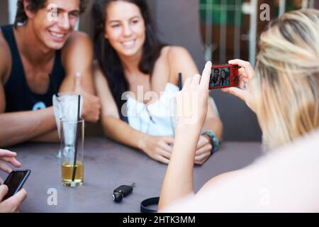 Immortalisez leurs plus beaux souvenirs. Groupe d'adolescents qui prennent des boissons dans un restaurant en plein air tout en prenant une photo avec un téléphone portable. Banque D'Images