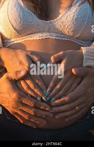 Le mari attentionné tenant le ventre de sa femme enceinte et tenant les mains de ses femmes pendant qu'elle fait une forme de coeur avec ses mains. Banque D'Images