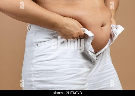 Femme avec grand ventre en flabby essayant de fermer les jeans blancs sur fond beige. Graisse viscérale. Corps positif. Gain de poids soudain. Petits vêtements serrés Banque D'Images