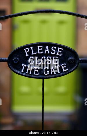 Une fonte métallique décorative audacieuse Veuillez fermer le panneau de la porte attaché à une porte en métal à l'extrémité d'un chemin de jardin à une maison avec une porte verte brillante. Banque D'Images