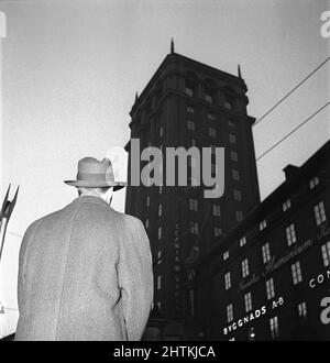 Dans le 1950s. Un homme dans un manteau et un chapeau est photographié dans la rue d'en dessous avec la silhouette d'un bâtiment haut en arrière-plan. Suède 1951 Kristoffersson réf. BE86-5 Banque D'Images