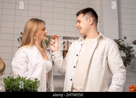 Couple heureux de cuisiner ensemble à la maison. Homme nourrissant petite amie ou femme tout en faisant des légumes frais salade dans la cuisine blanche moderne dans le style scani. Concept de week-end familial joyeux. Photo de haute qualité Banque D'Images