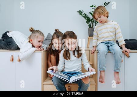 Des enfants s'ennuient se sont rassemblés dans une chambre pour lire un livre d'images, tous regardant. De cinq à huit ans. Banque D'Images