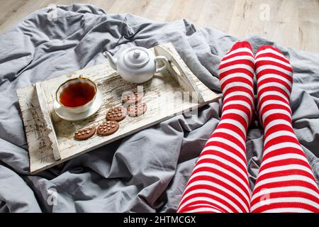 Fragments de jambes dans des chaussettes à rayures rouges. Concept intérieur et intérieur fantaisie. Vue de dessus. Une tasse de thé, une théière avec tisane, un bol à sucre sur une wh en bois Banque D'Images