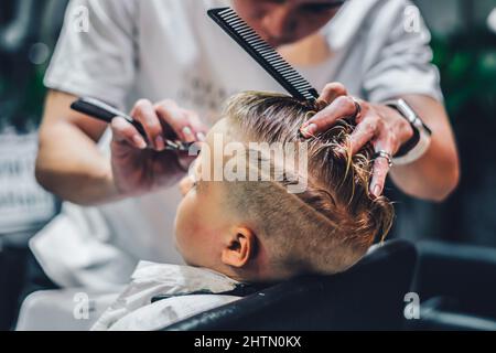 Style de vie photo salon de coiffure blond garçon client. Coiffeur homme travail déplacer main peigne outil ciseaux sèche-cheveux pince cape blanc. Cheveux courts enfants de style moderne Banque D'Images