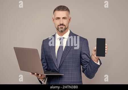 homme d'affaires confiant dans un costume professionnel tient un ordinateur portable et présente smartphone, publicité Banque D'Images