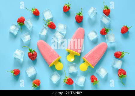 Pop-sicles faits maison. Crème glacée naturelle dans des moules en plastique brillant, des fraises et des glaçons sur fond bleu. Vue de dessus Flat lay. Banque D'Images