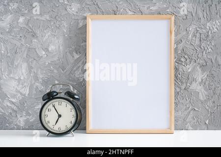 Cadre vide vertical en bois blanc et réveil noir sur table sur fond de mur en béton gris. Modèle de maquette pour votre conception. Banque D'Images
