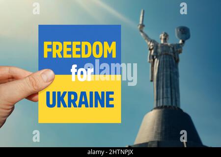 Main masculine tenant la carte drapeau de l'Ukraine avec le message de la liberté pour l'Ukraine devant le monument flou de la Terre mère à Kiev. Banque D'Images