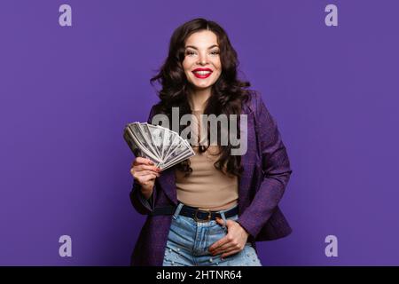 Une femme élégante montre un fan d'argent dans des billets de cent dollars sur fond violet. Fille dans le costume d'affaires tient beaucoup d'argent de papier d'argent liquide. Business, finance, épargne, banque et personnes concept Banque D'Images