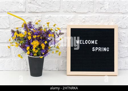 Texte Hello Spring sur tableau noir et bouquet de fleurs colorées en papier noir tasse à café avec paille cocktail sur fond blanc mur de brique. C Banque D'Images