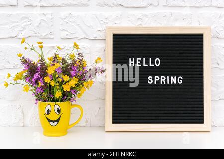 Texte Hello Spring sur le tableau de lettres noir et le bouquet de fleurs de couleur dans une tasse jaune drôle sur le fond blanc mur de brique. Concept Springtime humeur et Banque D'Images