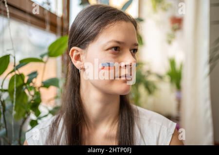 Une jeune fille russe triste avec le drapeau de l'Ukraine sur son visage regarde à travers la fenêtre. Le concept de la participation du peuple ukrainien au Banque D'Images