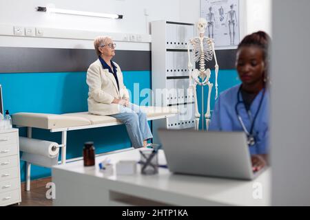 Infirmière spécialisée travaillant dans le traitement de santé dactylographiant l'expertise de la maladie sur ordinateur alors que la patiente âgée debout sur le lit attendant le diagnostic de la maladie dans le bureau de l'hôpital. Assistance technique Banque D'Images