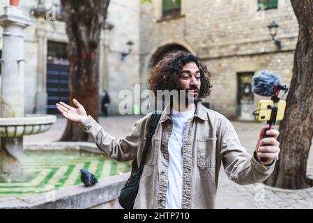 Blogueur mâle barbu enregistrant la vidéo sur un smartphone moderne avec microphone tout en faisant des gestes dans la rue près d'une fontaine en ville pendant le voyage Banque D'Images