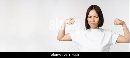 Femme asiatique souriante montrant des biceps flexibles, des muscles forts bras gestuelle, debout dans un t-shirt blanc sur fond blanc Banque D'Images