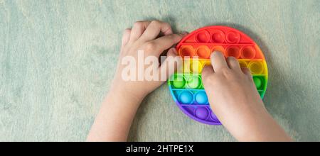 Bannière avec enfant jouant avec le jouet sensoriel pop IT. Garçon appuyant sur coloré arc-en-ciel squishy doux bulles de silicone. Soulagement du stress et de l'anxiété. Jeu de violon tendance. Vue du dessus Banque D'Images