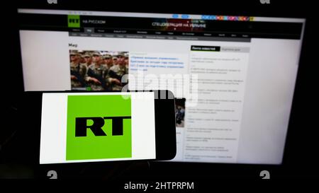 Personne tenant un téléphone portable avec le logo de la société russe de télévision contrôlée par l'État RT à l'écran devant la page Web. Mise au point sur l'affichage du téléphone. Banque D'Images