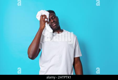 Un jeune homme afro-américain essuye la transpiration de son visage avec une serviette blanche sur un fond bleu isolé après un entraînement. Un mode de vie sain Banque D'Images